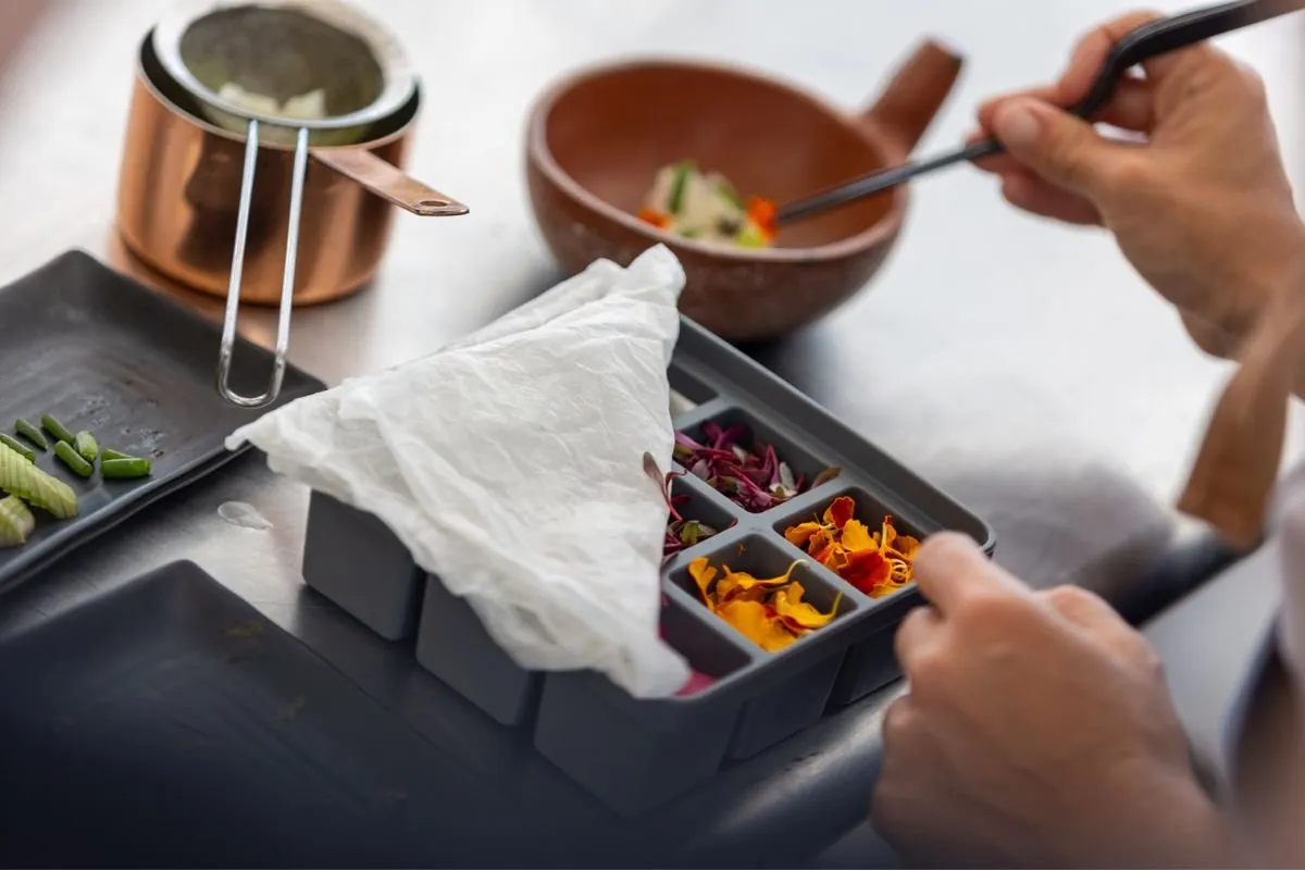 Hands selecting herbs and flowers with tweezers from a compartmentalised tray