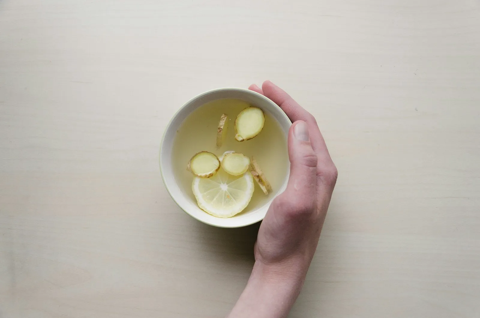 A hand holds a white bowl with slices of lime and ginger in water