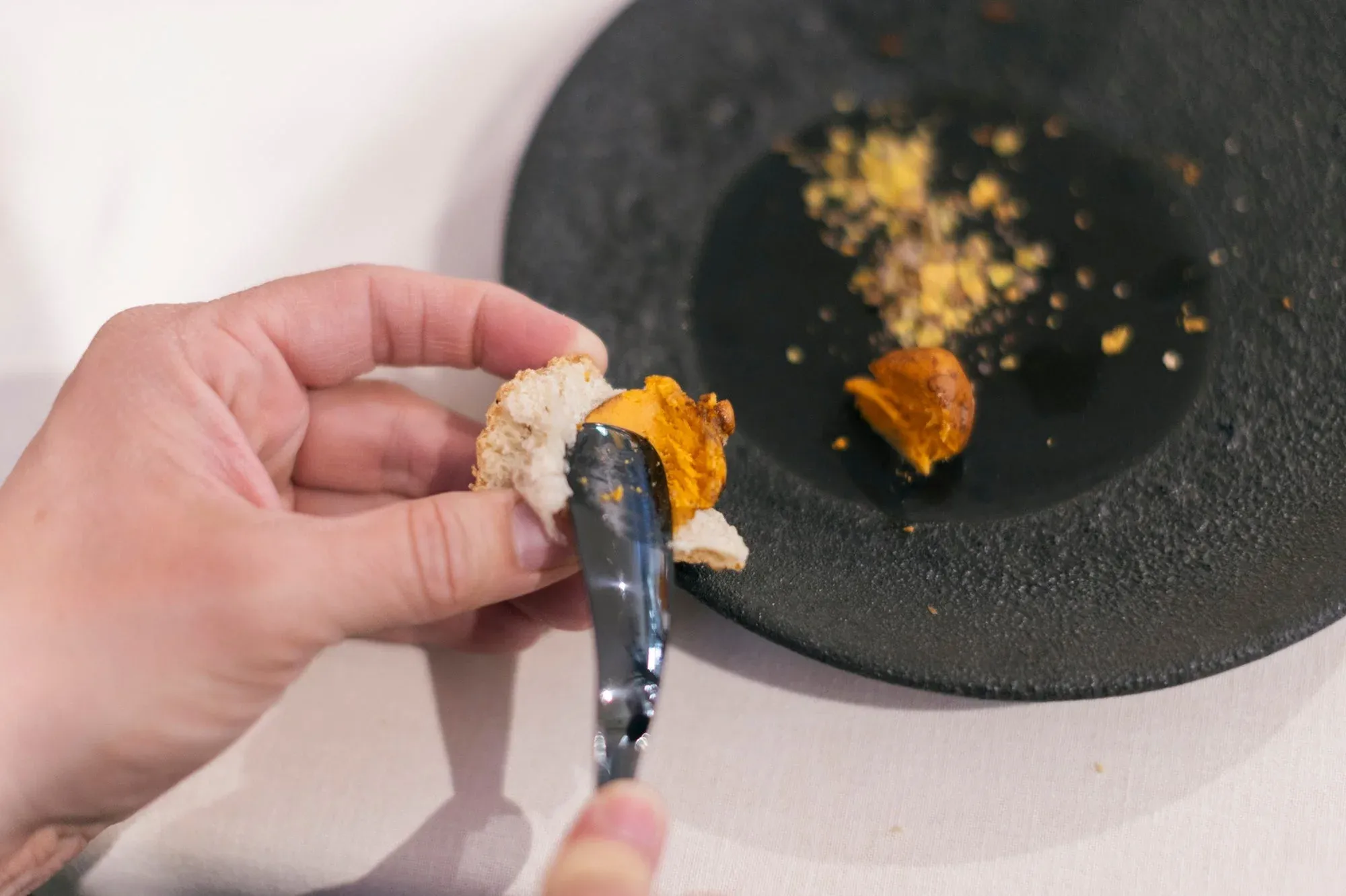 Una mano unta un bocado sobre un plato de piedra negra con migas y una crema anaranjada