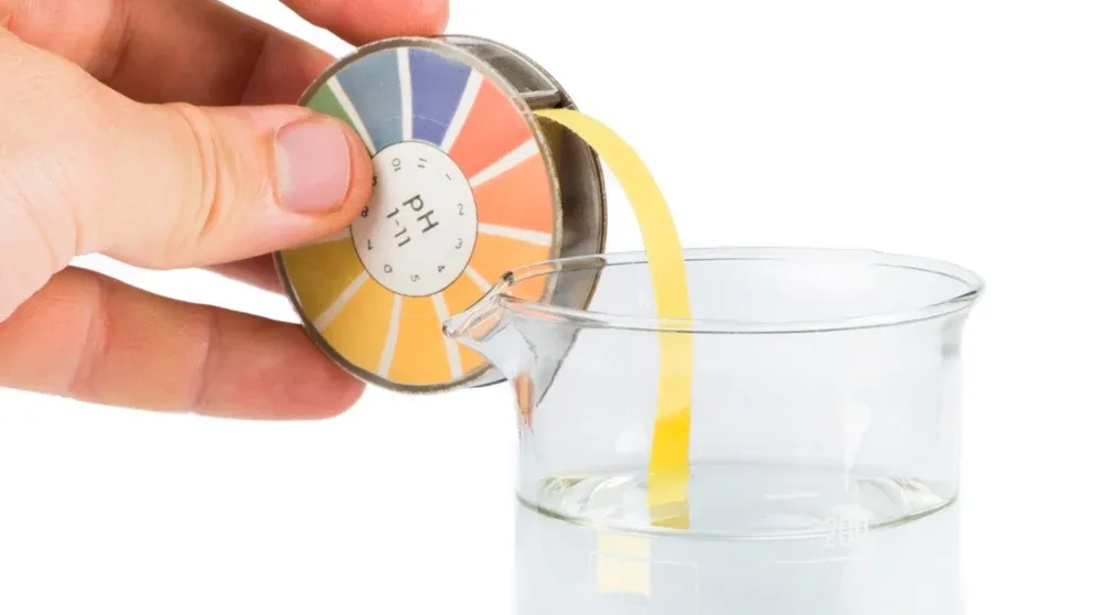 A hand holds a pH colour chart next to a beaker with liquid