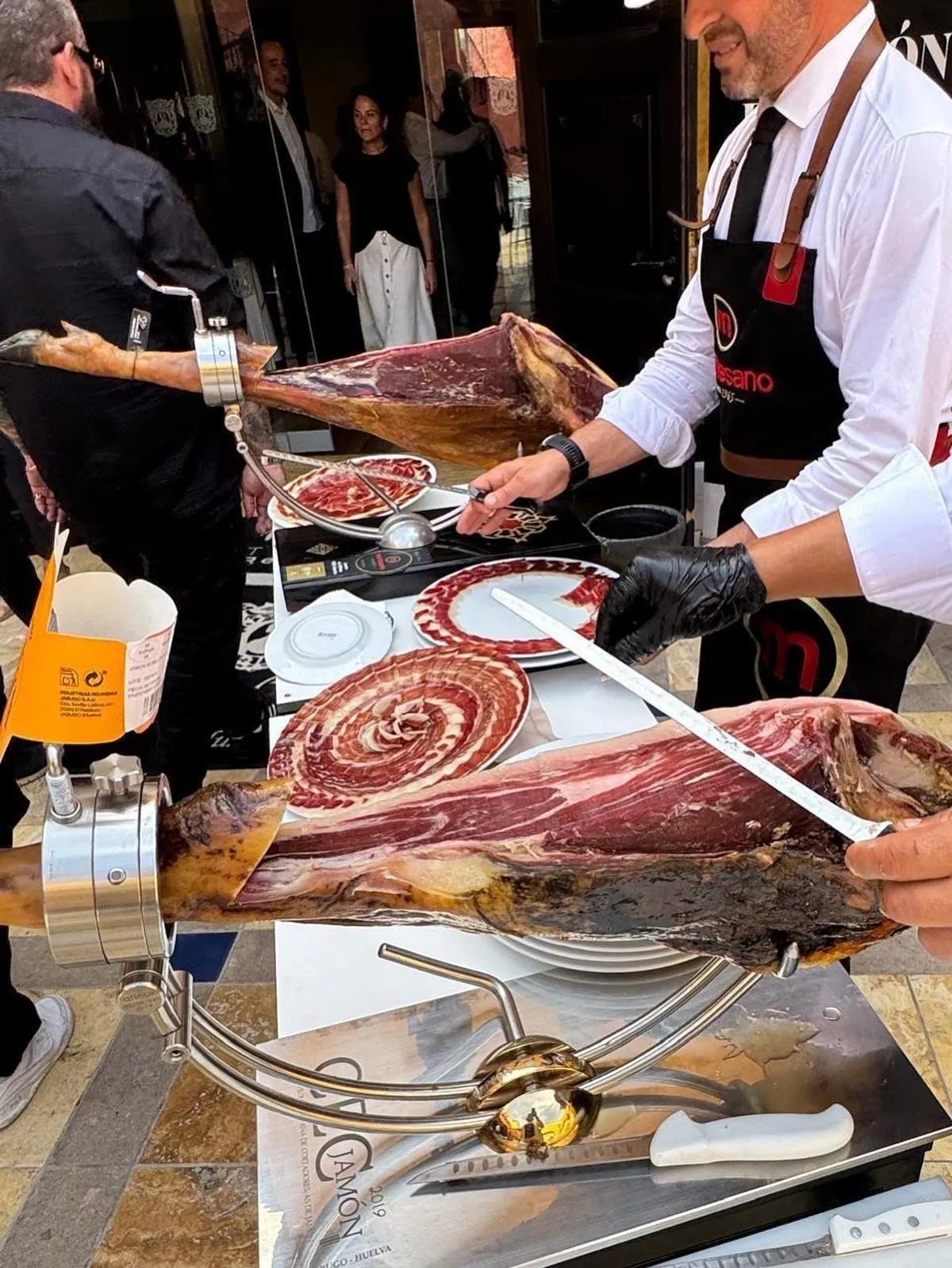 Corte de jamón a pie de entrada del restaurante: un cortador con delantal de Montesano y guante negro desliza el cuchillo jamonero sobre la pieza, con un segundo jamón montado en otro jamonero al lado y varios platos ya montados con las lonchas en espiral