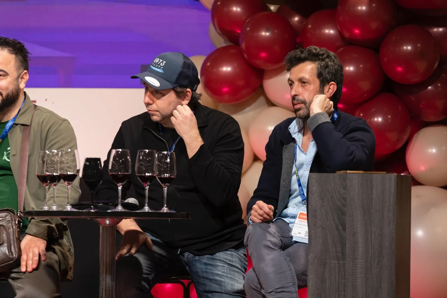 Diego Schattenhofer, wearing his Taste 1973 cap, seated in front of a row of red wine glasses during a congress tasting, with maroon and cream balloons in the background