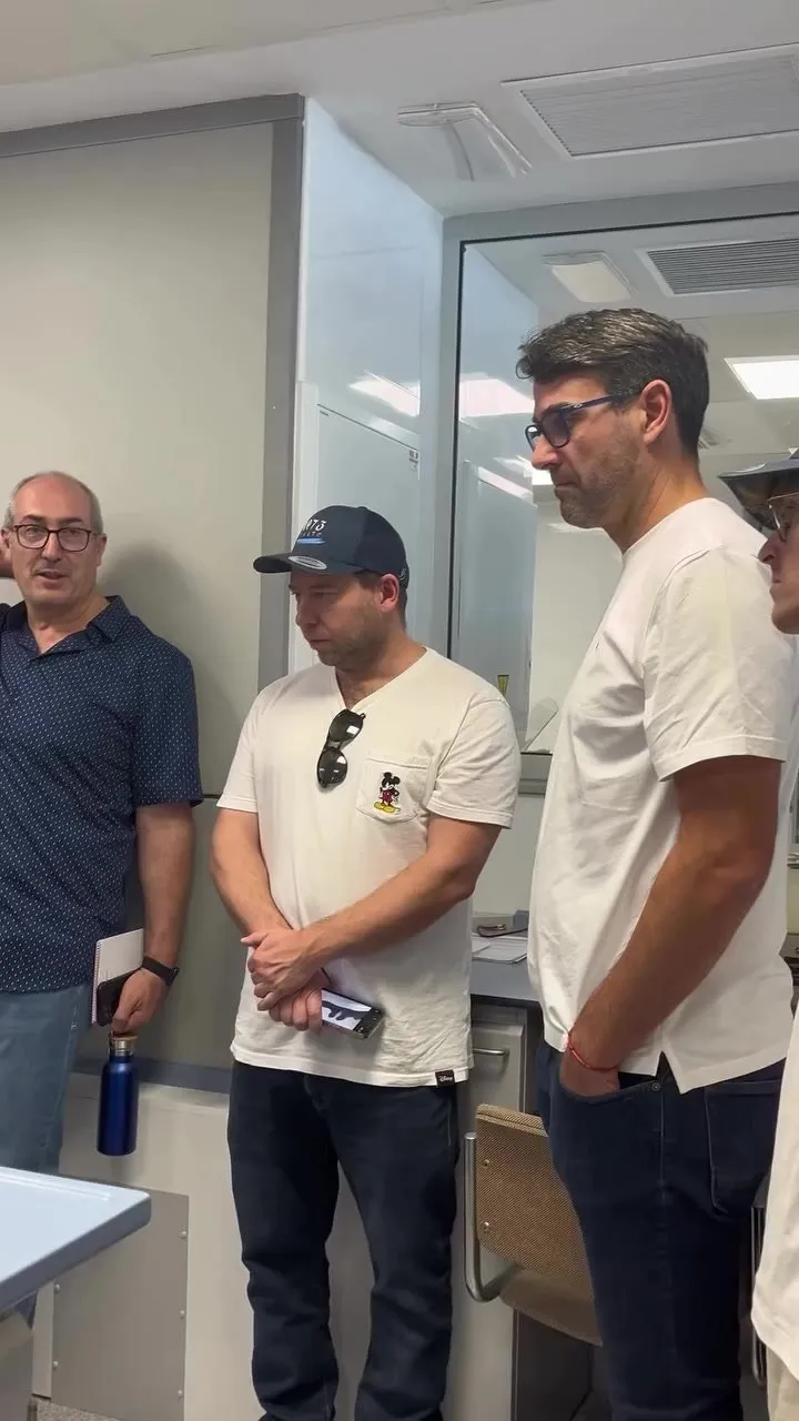 Four people stand in a light-walled laboratory listening attentively; among them, a man in a dark 1973 cap and white T-shirt with his sunglasses hanging from his neck