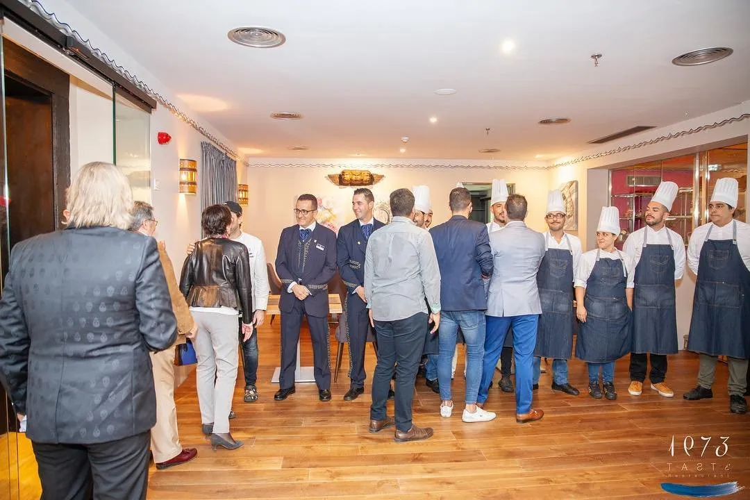 Equipo de cocina con gorros y delantales vaqueros, en fila junto al personal de sala del hotel, charlando con los invitados en la sala recién terminada