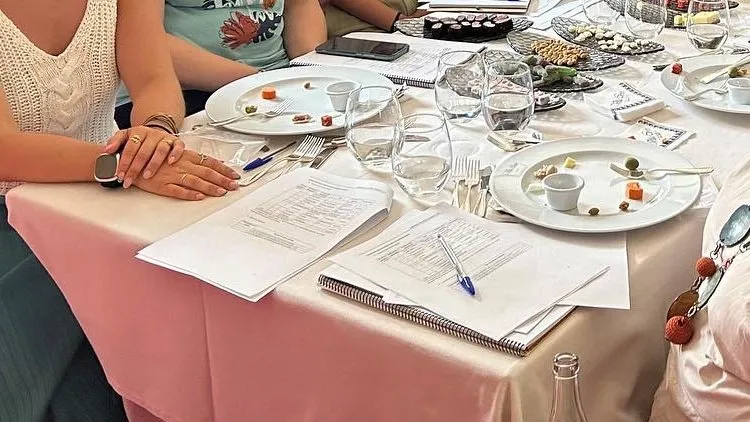 Detail of the workshop table: two white plates with small bites of meat, green olives and a small white bowl, glasses and tumblers, and on the tablecloth, printed tasting sheets and a spiral notebook with a pen on top