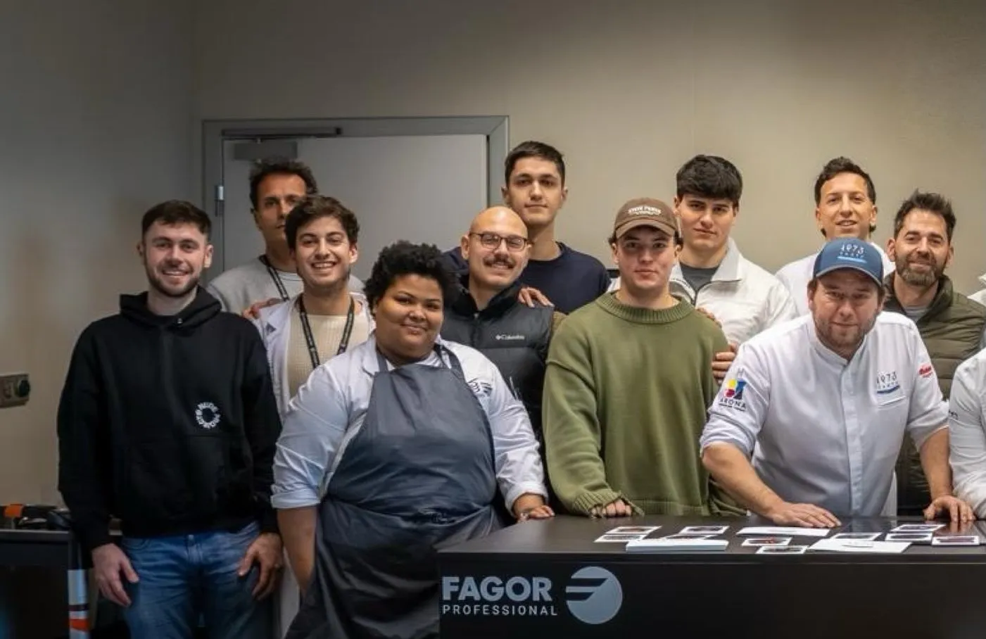A dozen people, chefs in jackets and attendees in street clothes, pose together behind a work table bearing the sign “Fagor Professional”; on the right, Diego Schattenhofer wearing a blue cap and Taste 1973 chef’s jacket