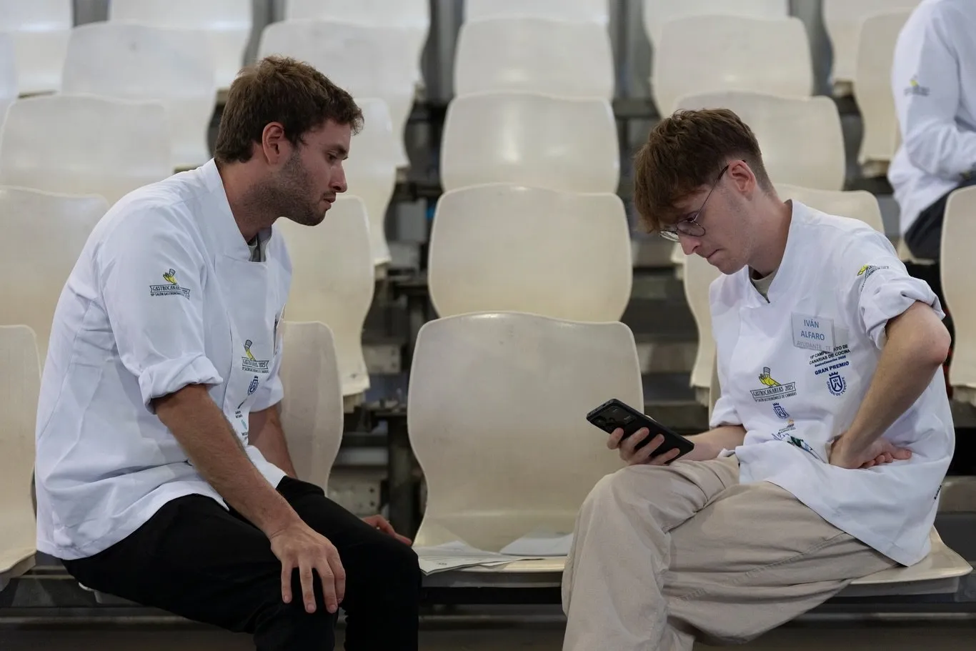 Two chefs in white jackets sitting in the stands of the fairground; the one on the right has printed on his chest the accreditation of Iván Alfaro as assistant of the 19th Canary Islands Cooking Championship