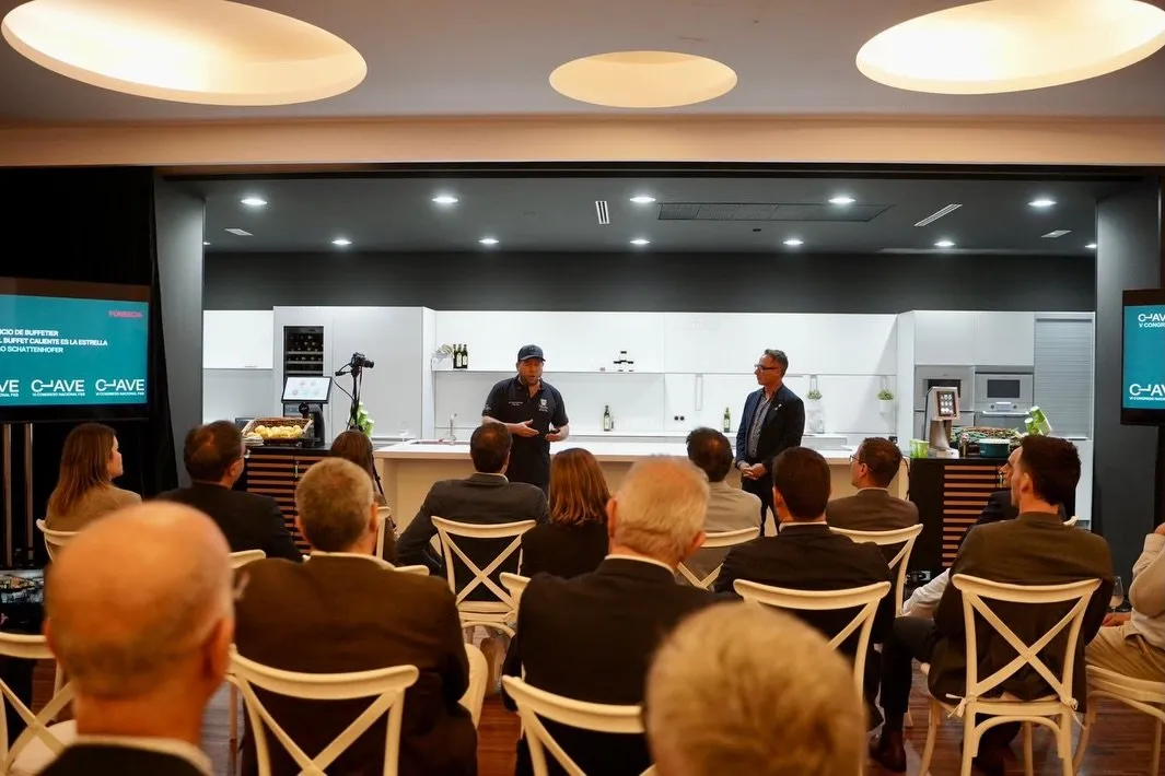 Sala del congreso con el público sentado en sillas blancas frente a una cocina de exposición iluminada, donde Diego Schattenhofer, con gorra oscura y micrófono de diadema, interviene de pie junto a un hombre de traje; en las pantallas laterales se ven el logotipo del congreso y el rótulo de la ponencia
