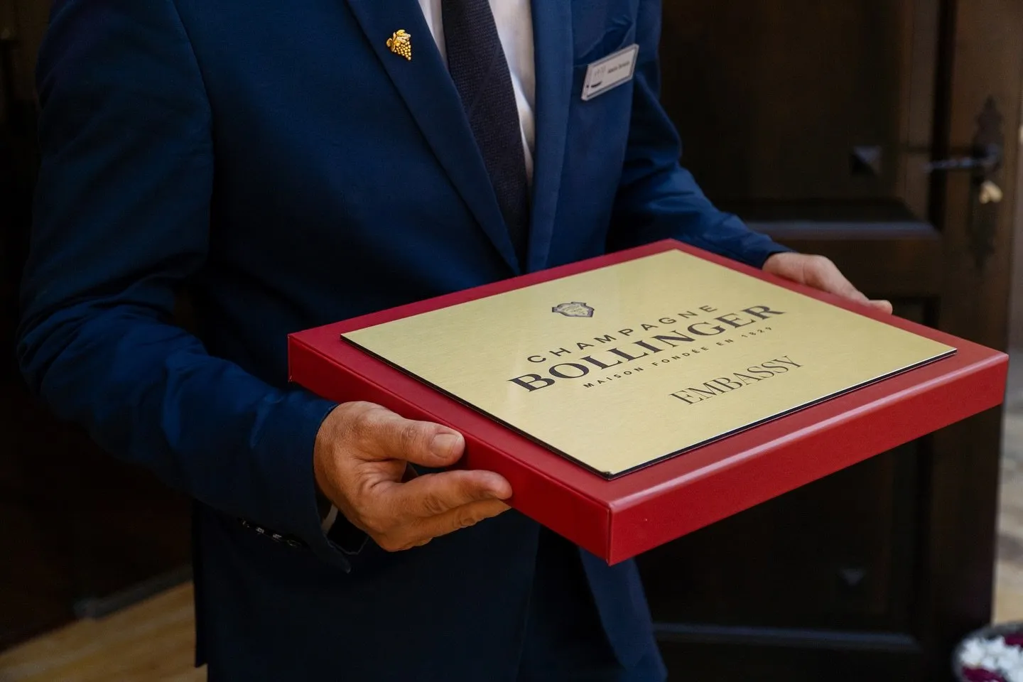 Close-up shot of a man in a navy-blue suit, with a golden bunch of grapes on his lapel, holding with both hands a golden plaque set in its red presentation box, with the house’s crest and the engraved text Champagne Bollinger, Maison Fondée en 1829, Embassy