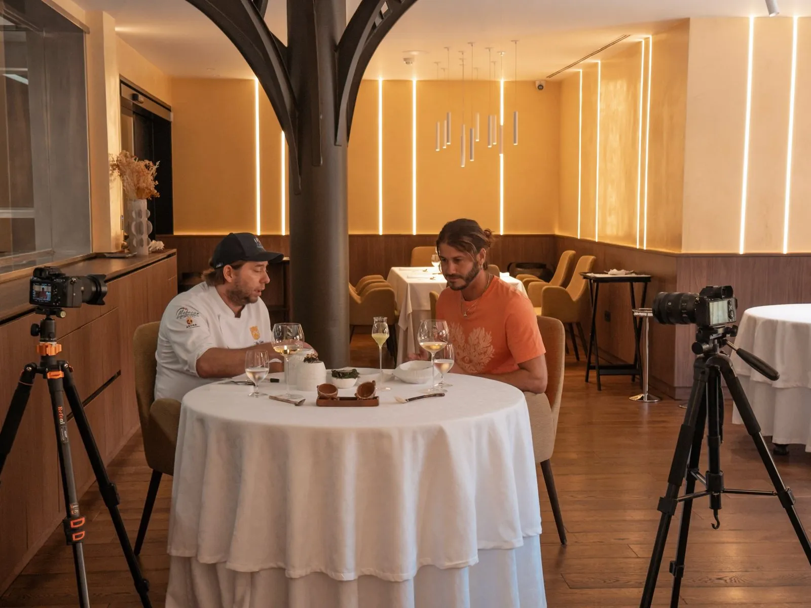 Diego Schattenhofer y un miembro del equipo de «Ecos del Mar» sentados a una mesa redonda del comedor de Taste 1973, con dos trípodes de cámara enfocándolos desde ambos lados y el árbol escultórico de la sala al fondo