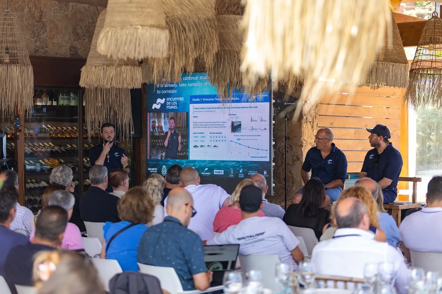 General view of the panel room: a speaker standing next to a screen showing a slide on temporal variations of metals, with two panellists seated to the right in front of a large audience