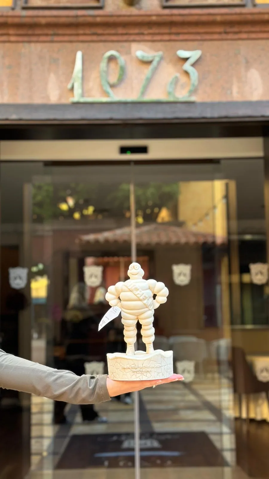 A hand holds the white Michelin Man trophy figure, its base engraved with the Barcelona gala, in front of the restaurant’s entrance door marked with the number 1973