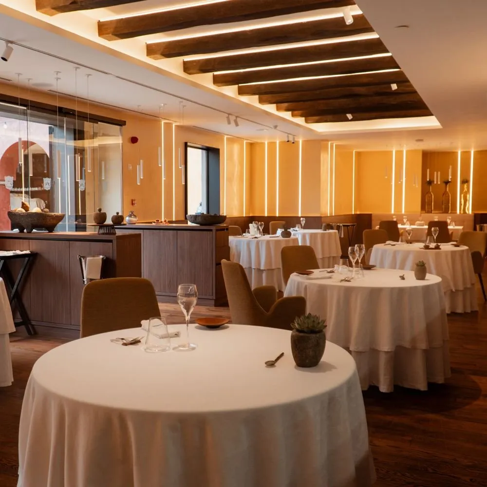 Sala de Taste 1973 con las mesas redondas vestidas de blanco, las vigas de madera del techo y líneas de luz cálida sobre la pared ocre