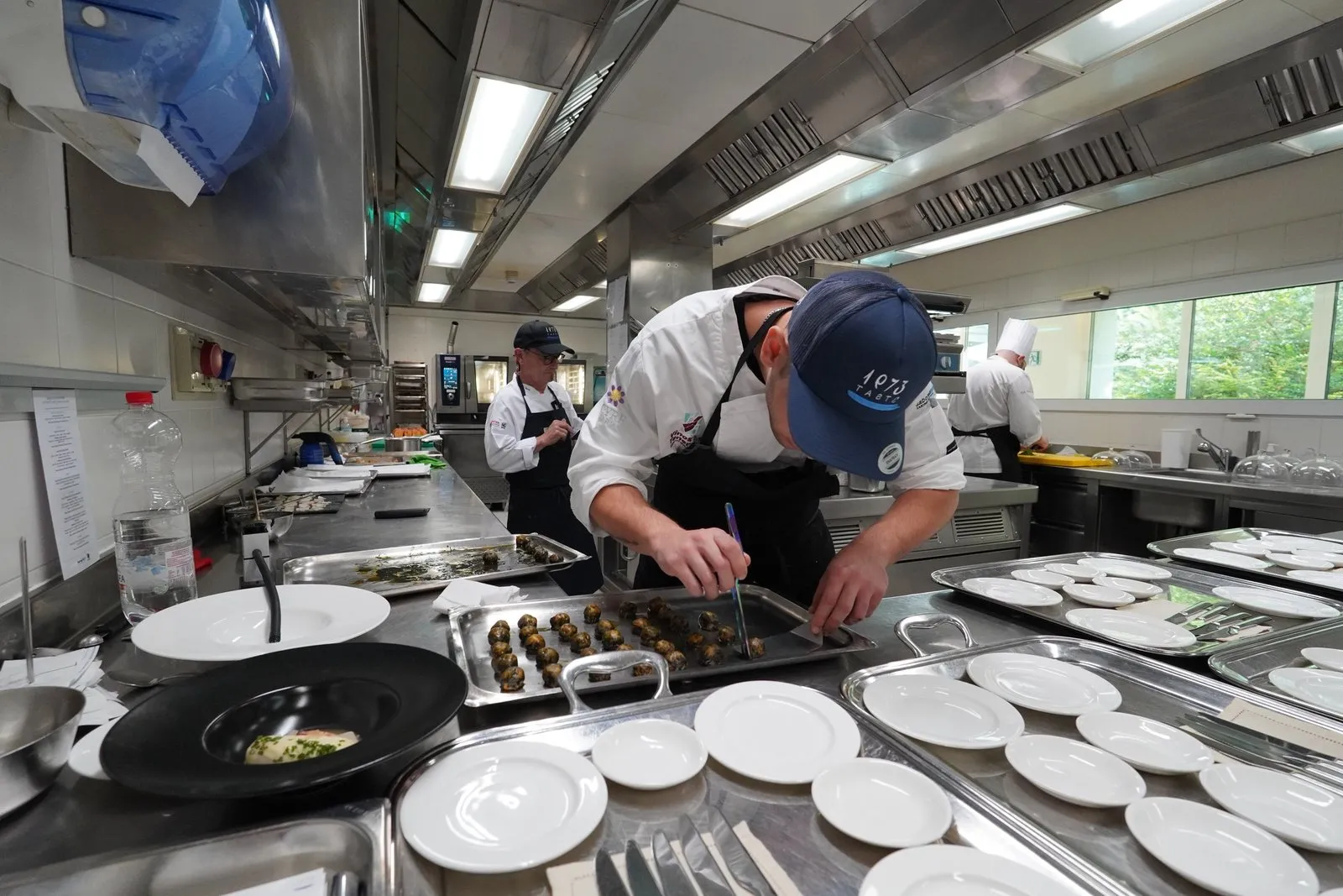 Diego Schattenhofer, con la gorra de Taste 1973, coloca con unas pinzas los bocados de una bandeja de acero en el pase de la cocina, rodeado de filas de platos vacíos