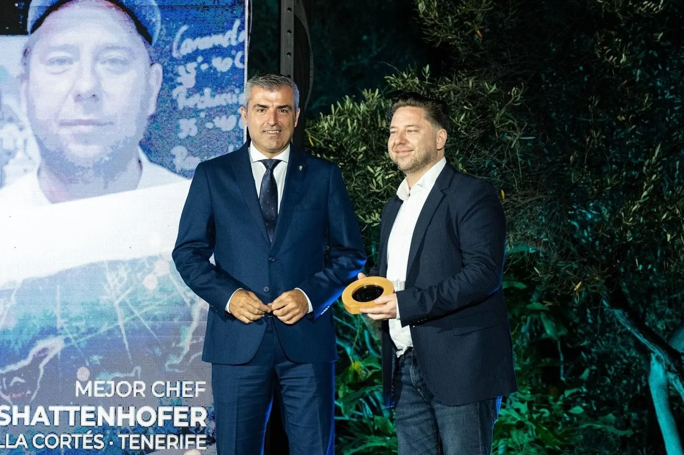 Diego Schattenhofer recibe el galardón en el escenario junto a un hombre de traje azul, con su retrato y el rótulo «Mejor chef» proyectados en la pantalla del fondo