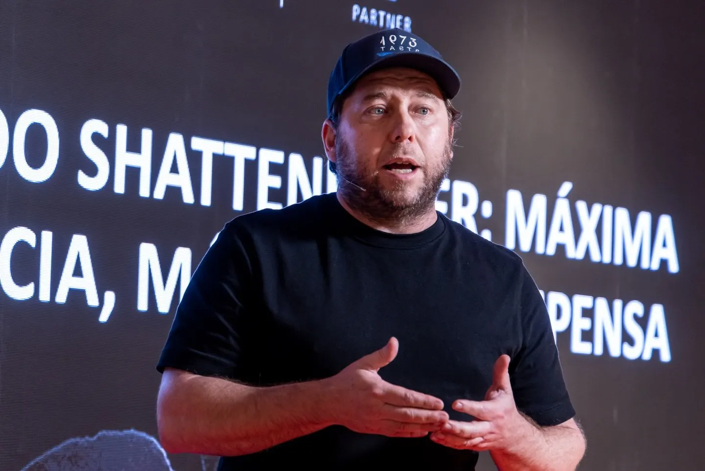 Diego Schattenhofer, con gorra de 1973 Taste y camiseta negra, habla con micrófono de diadema ante una pantalla con su apellido proyectado