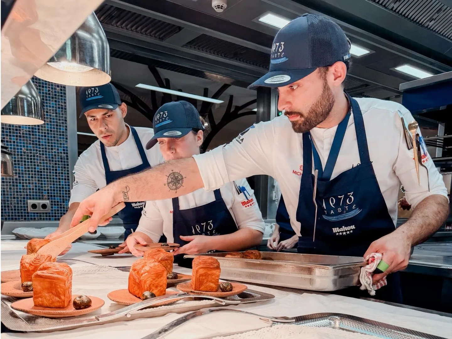 Tres cocineros con gorra y mandil de Taste 1973 rematan en el pase una fila de bocados hojaldrados servidos sobre platos de barro