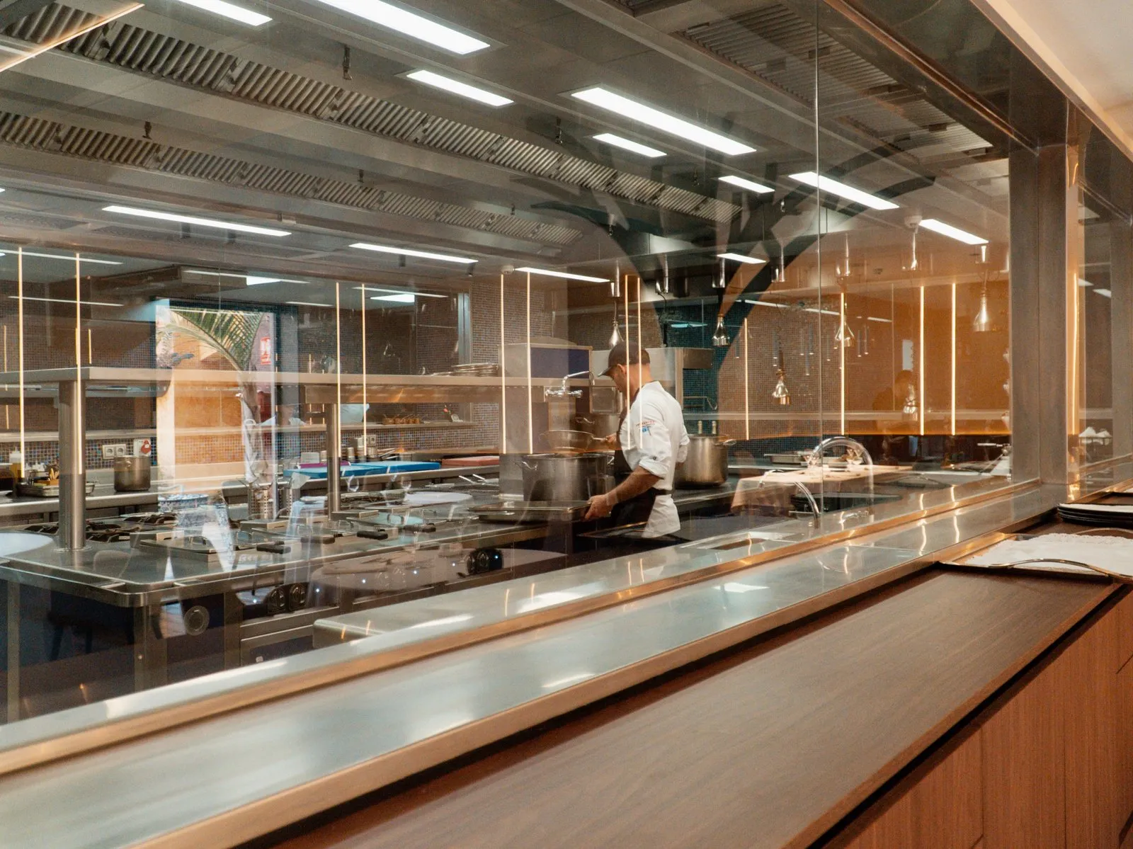 Un cocinero trabaja entre cazuelas en los fogones de Taste 1973, visto desde el comedor a través del cristal de la cocina vista