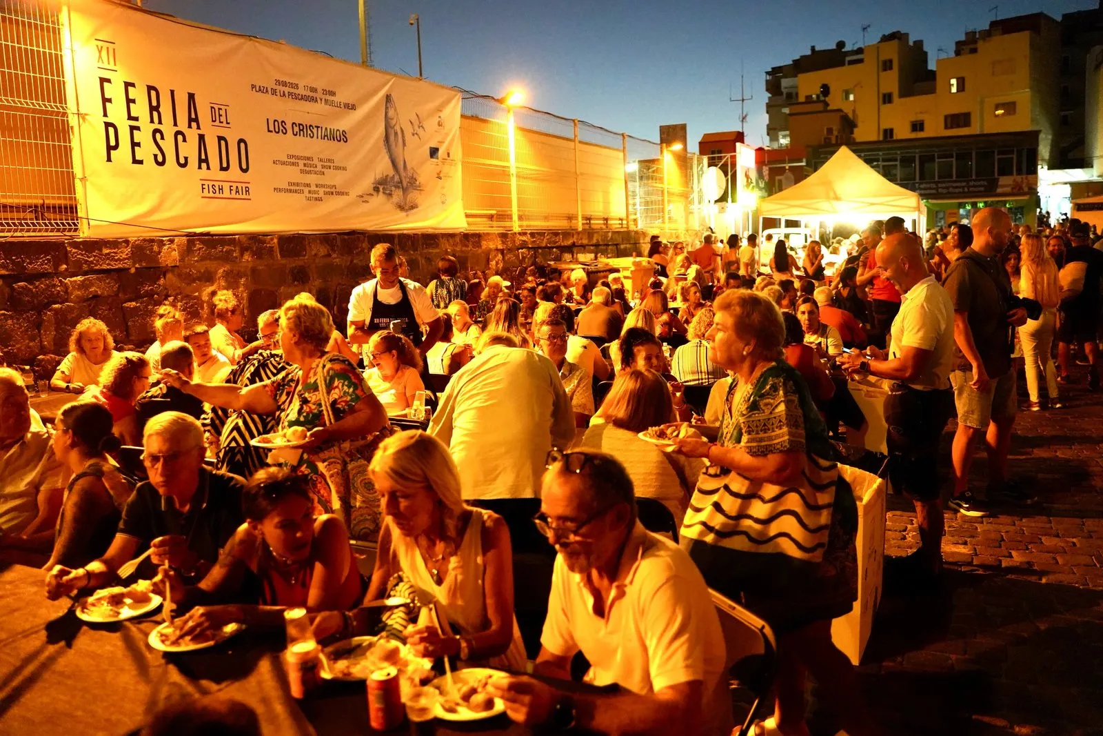 Cientos de personas cenan en mesas corridas al anochecer junto a una lona blanca que anuncia la XII Feria del Pescado de Los Cristianos