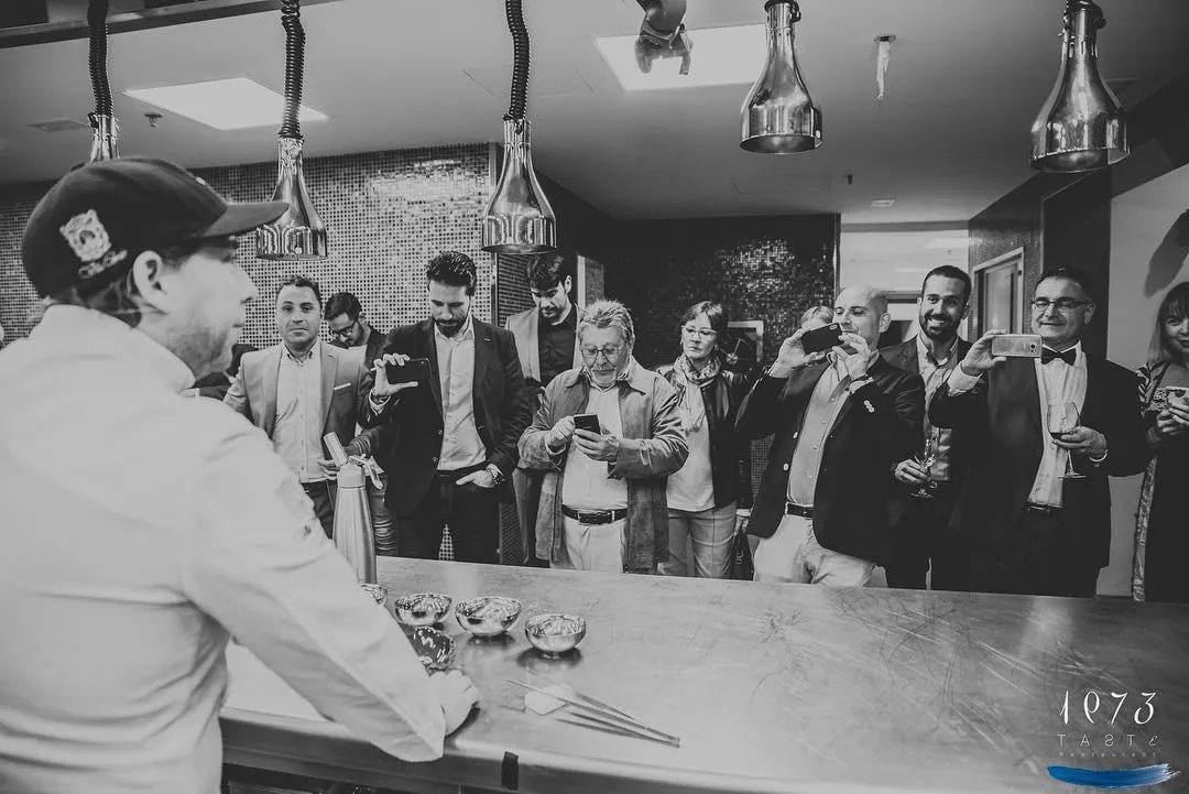 Fotografía en blanco y negro tras la barra de cocina abierta: un cocinero de espaldas presenta unos cuencos metálicos a los invitados, que graban con el móvil y sostienen copas de vino