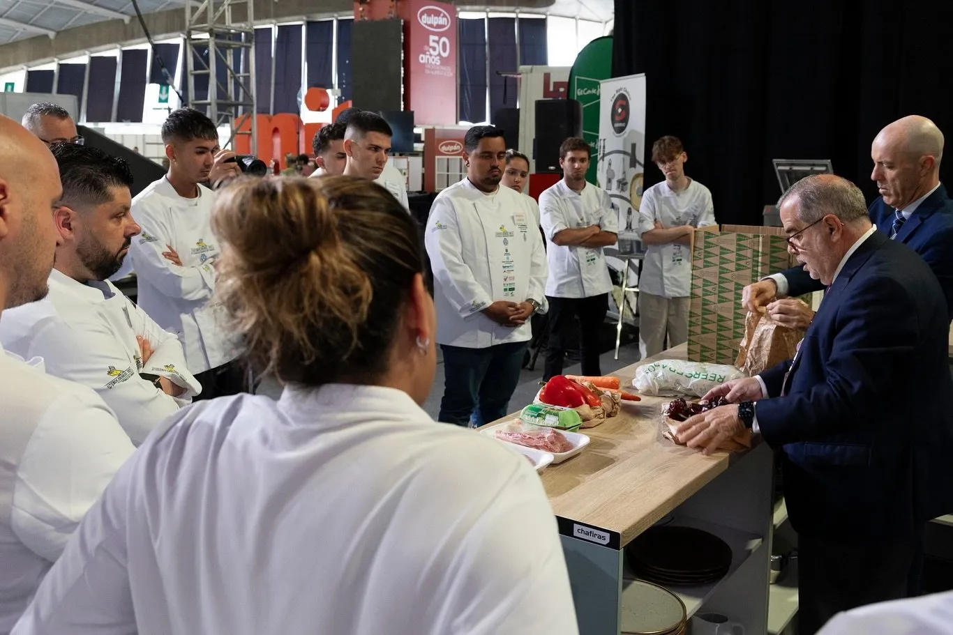 A jury member in a suit uncovers the ingredients of the surprise box on the work table while the competing chefs look on in a circle