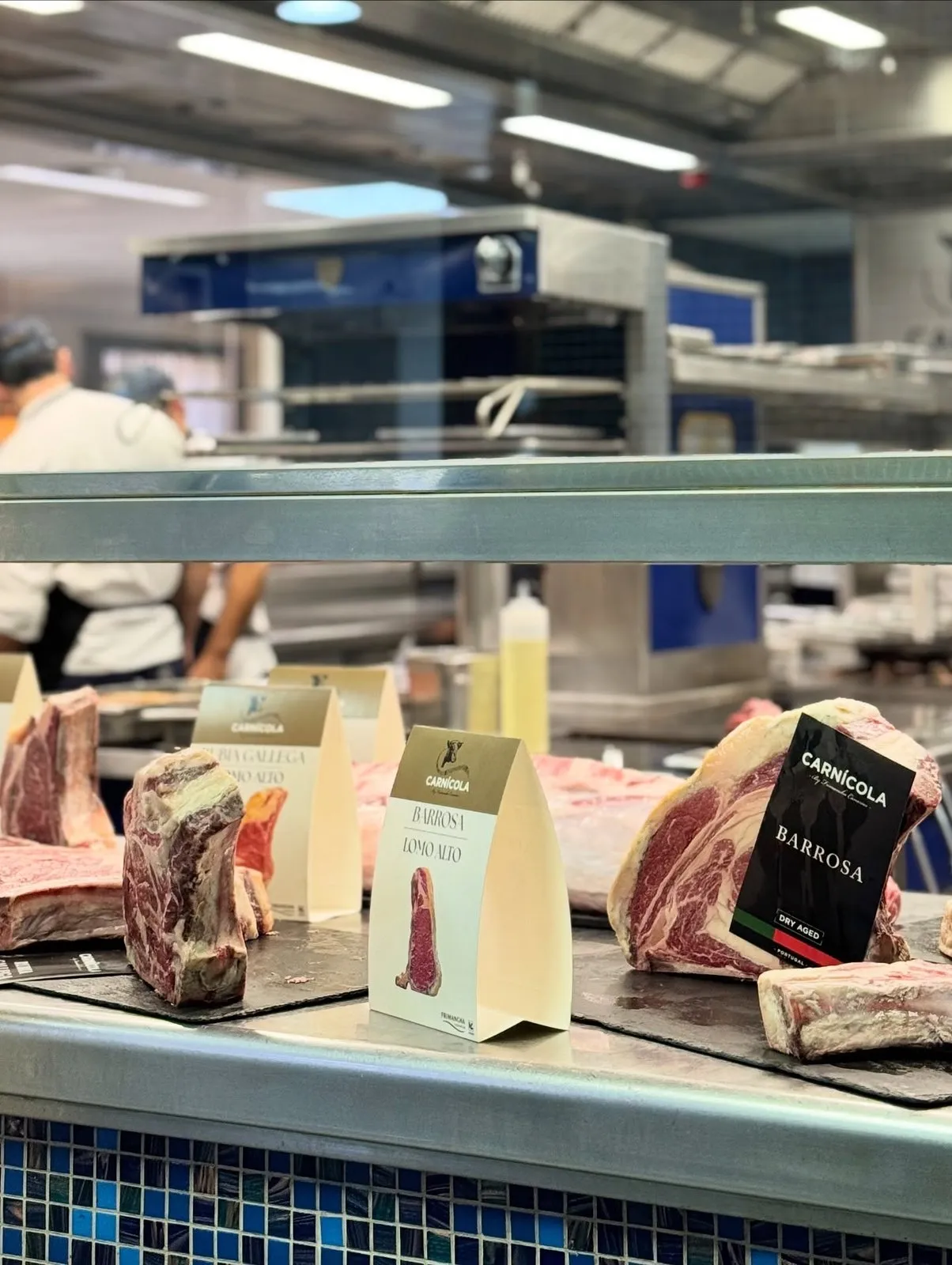 Mostrador de acero del economato con piezas de vacuno sobre bandejas de pizarra y carteles de la marca Carnícola que rotulan «Rubia Gallega Lomo Alto», «Barrosa Lomo Alto» y «Barrosa Dry Aged, Portugal»; al fondo, la cocina