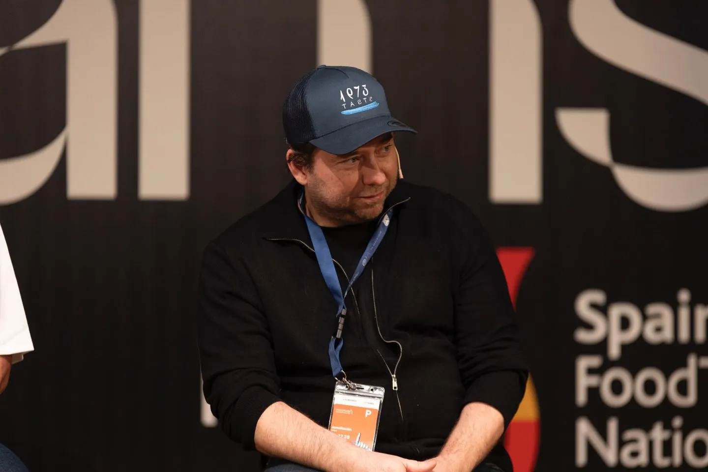 Diego Schattenhofer, wearing a navy Taste 1973 cap and an accreditation badge around his neck, listens as he sits on stage in front of the black Spain FoodTech Nation panel