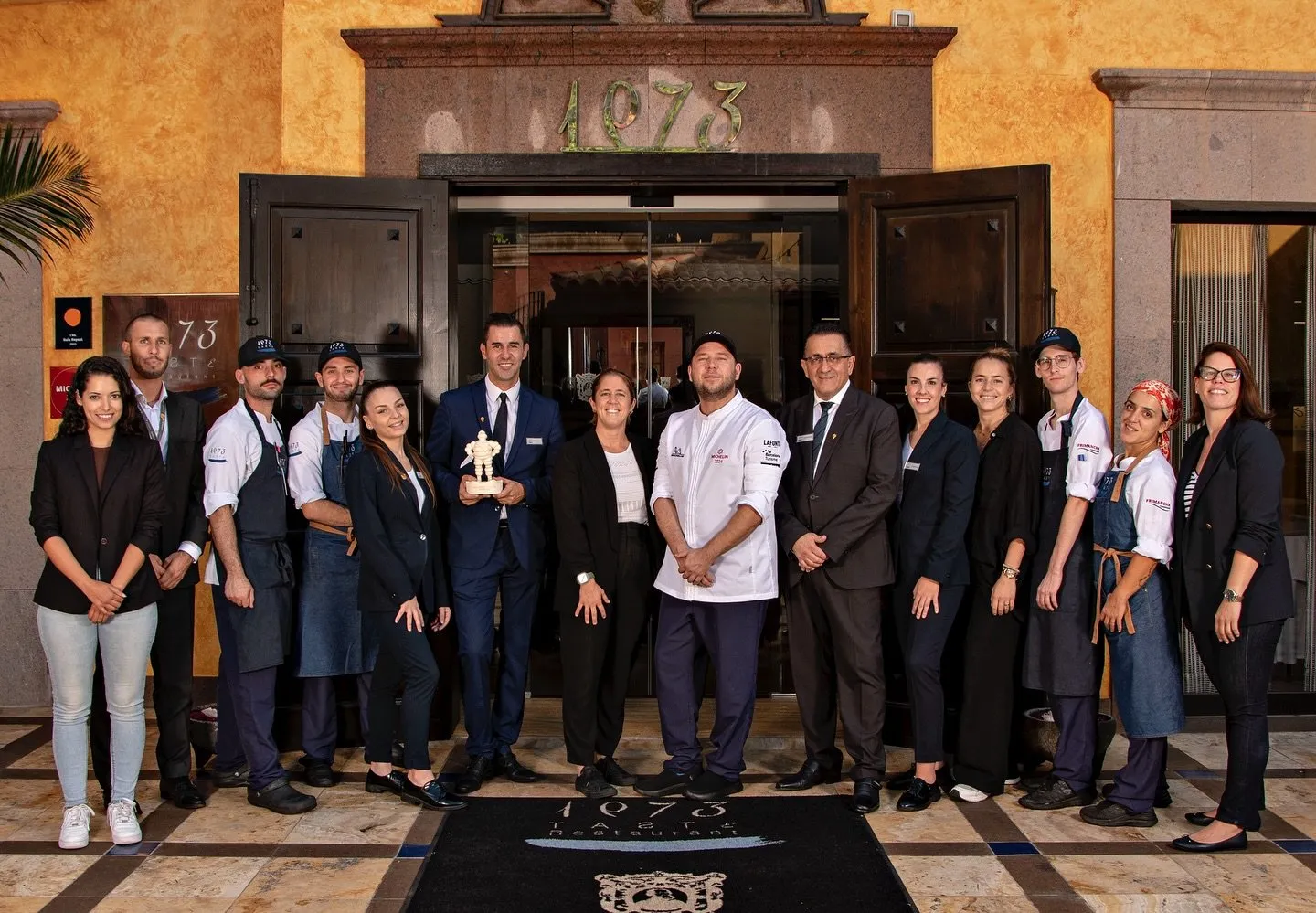 The full Taste 1973 team, kitchen and dining room, poses at the restaurant entrance with the Michelin trophy at the centre, beneath the 1973 sign