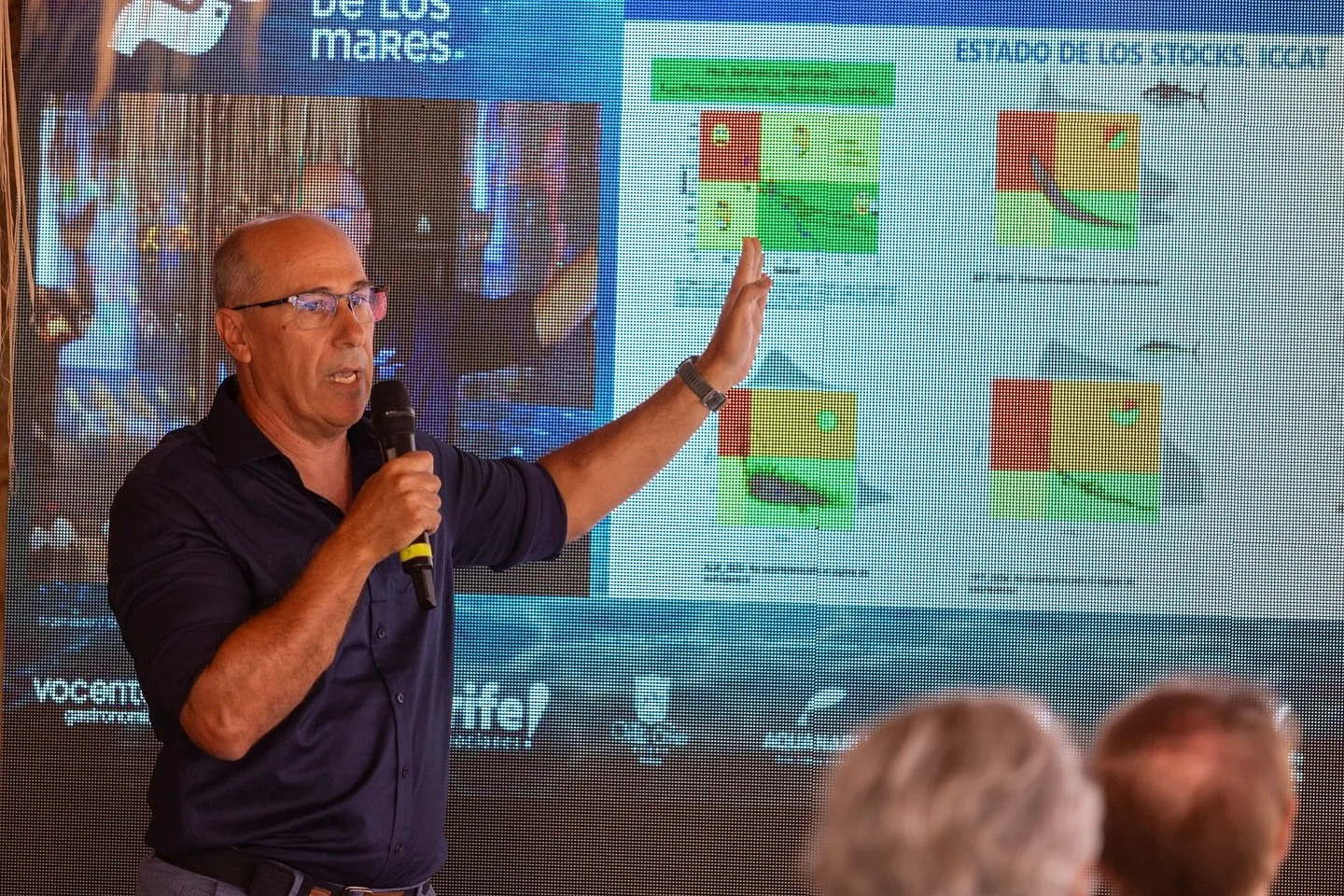 A researcher holding a microphone points to a screen with the Encuentro de los Mares logo and several coloured graphs under the heading “Estado de los stocks” (State of the stocks)