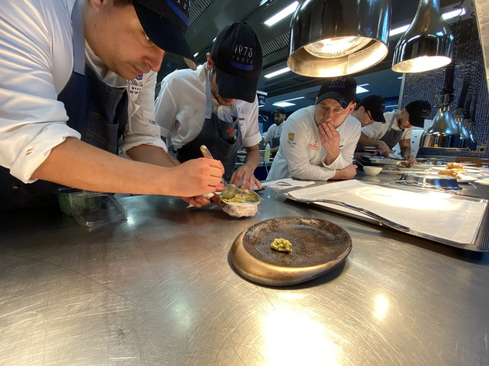 Several cooks wearing “1973 TASTE” caps bent over the steel pass under the heat lamps; one plates with a spoon while Diego Schattenhofer watches with his hand on his chin and, in the foreground, a golden ceramic plate waits