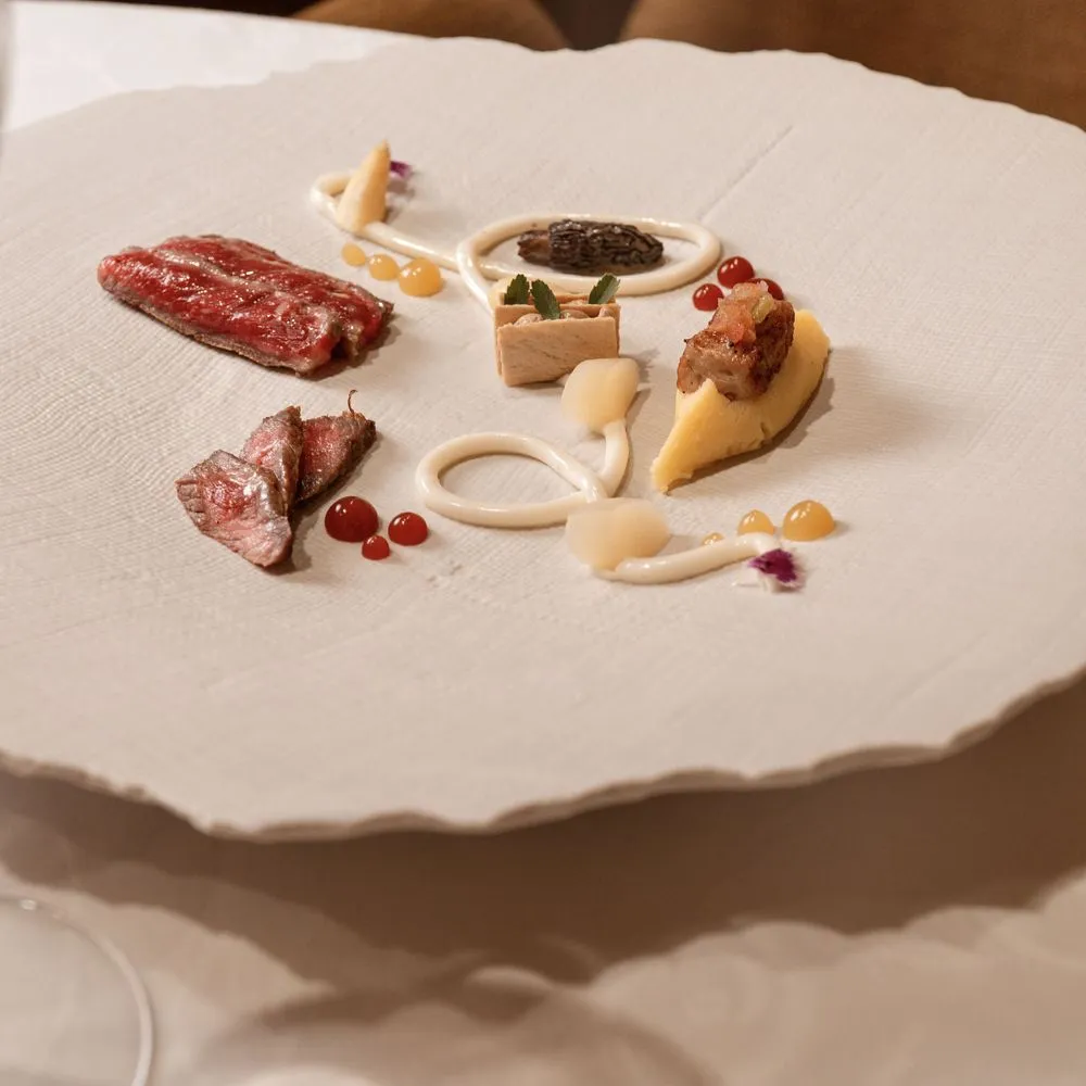 Plato blanco texturado con láminas de carne poco hecha, una morilla, puré y puntos de salsa