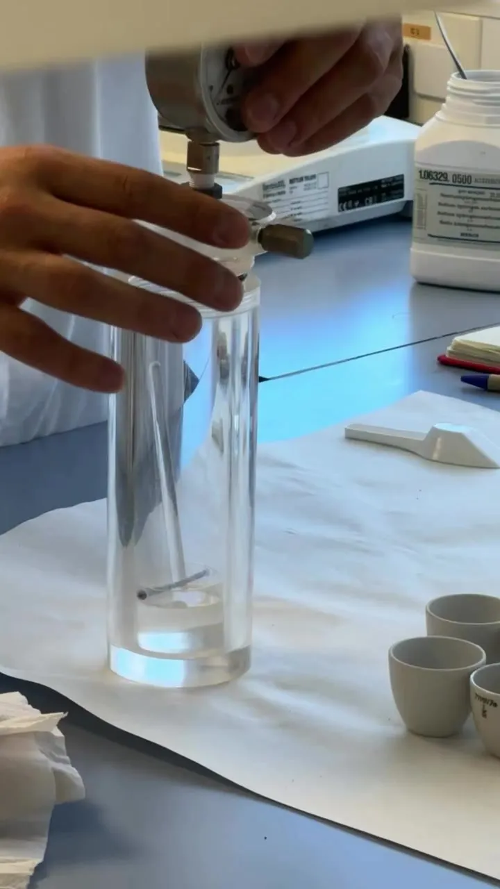 Two hands hold a transparent glass measuring cylinder with a small amount of liquid, beneath a manometer and next to a precision balance and several ceramic crucibles on a white cloth