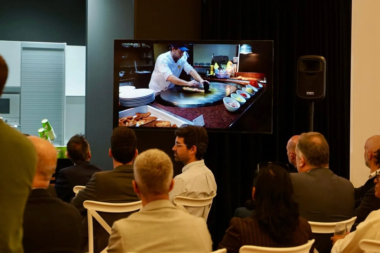 Pantalla grande en la sala del congreso con un vídeo de un cocinero trabajando sobre una plancha circular rodeada de cuencos con ingredientes, mientras el público sigue la proyección de espaldas a la cámara