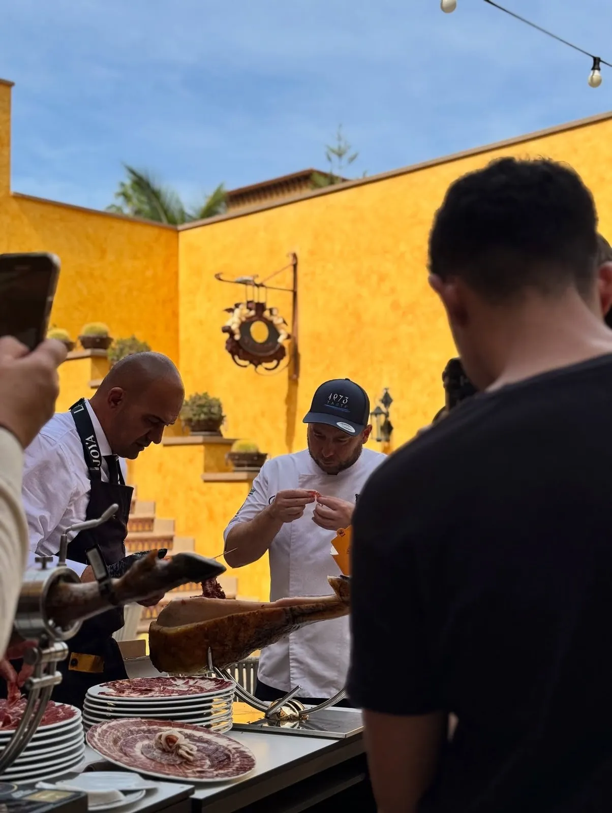 Diego Schattenhofer, con la gorra azul de 1973 Taste y chaquetilla blanca, sostiene con los dedos una loncha de jamón recién cortada y la observa; frente a él, un cortador con delantal de La Joya sigue trabajando la pieza sobre el jamonero