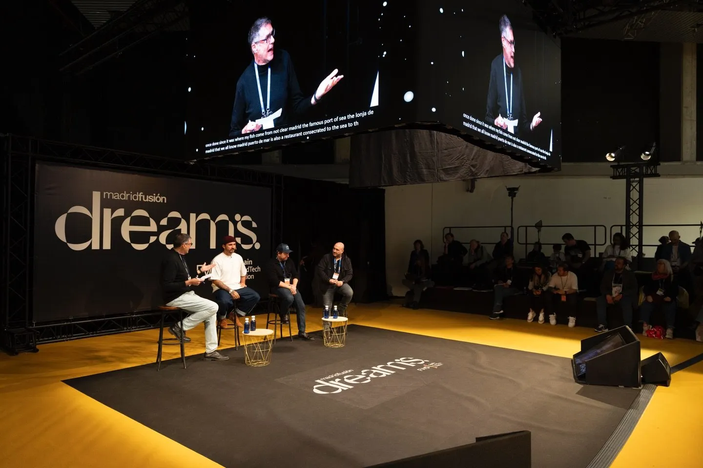 Wide shot of the Madrid Fusión Dreams stage during the round table, with the four participants on stools, the giant screen lit up above them and the audience seated to the right