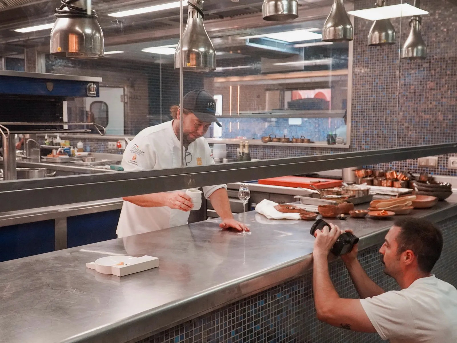 Diego Schattenhofer sirve y explica un plato desde la barra pasavante de la cocina de Taste 1973 mientras un cámara del equipo lo graba con la cámara en mano desde el otro lado