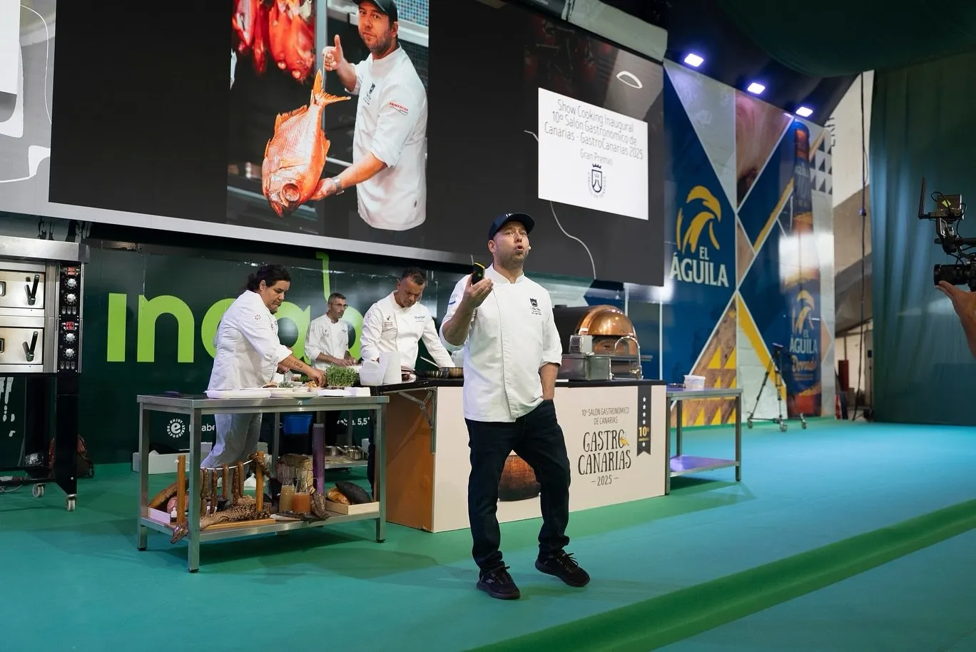 Diego Schattenhofer, wearing a cap and white chef’s jacket, speaks into a headset microphone at the lectern of the Opening Show Cooking of the 10th Salón Gastronómico de Canarias, with three chefs working behind him and a giant screen showing a chef holding a fish