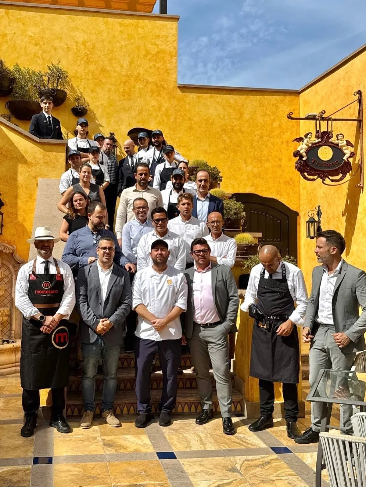 Una veintena de personas, cocina y sala de Taste 1973, posan escalonadas en la escalinata de la terraza del restaurante, con la pared ocre al fondo; en primera fila, dos cortadores con delantal de Montesano y de La Joya flanquean a Diego Schattenhofer y a varios representantes de los patrocinadores