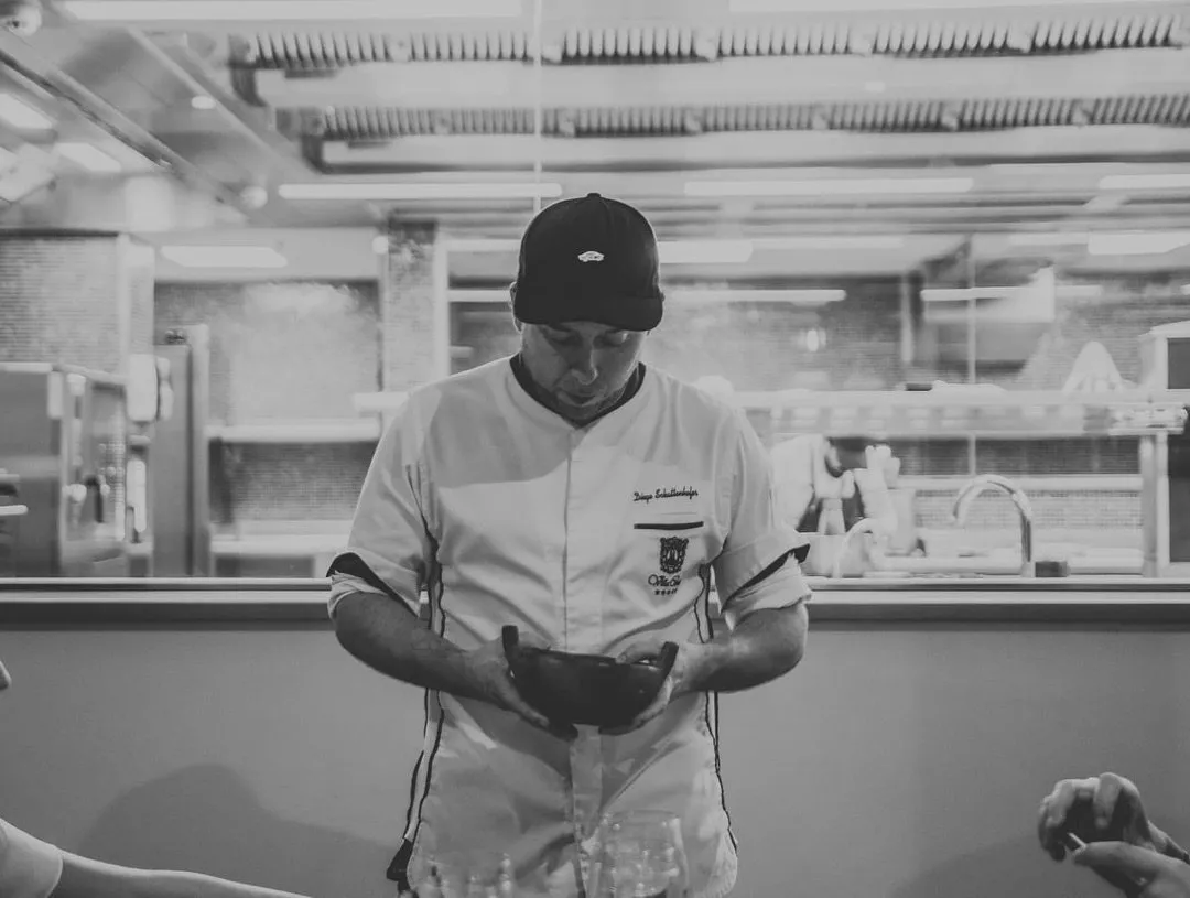 Fotografía en blanco y negro de Diego Schattenhofer, con gorra y chaquetilla bordada, de pie en la cocina y mirando el cuenco oscuro que sostiene con las dos manos