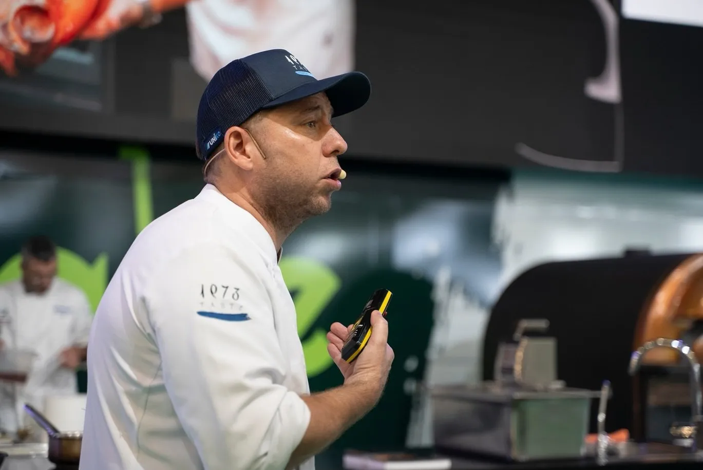 Close-up of Diego Schattenhofer in profile, wearing a cap and a chef’s jacket embroidered with the 1973 Taste logo, speaking into the headset microphone during his presentation