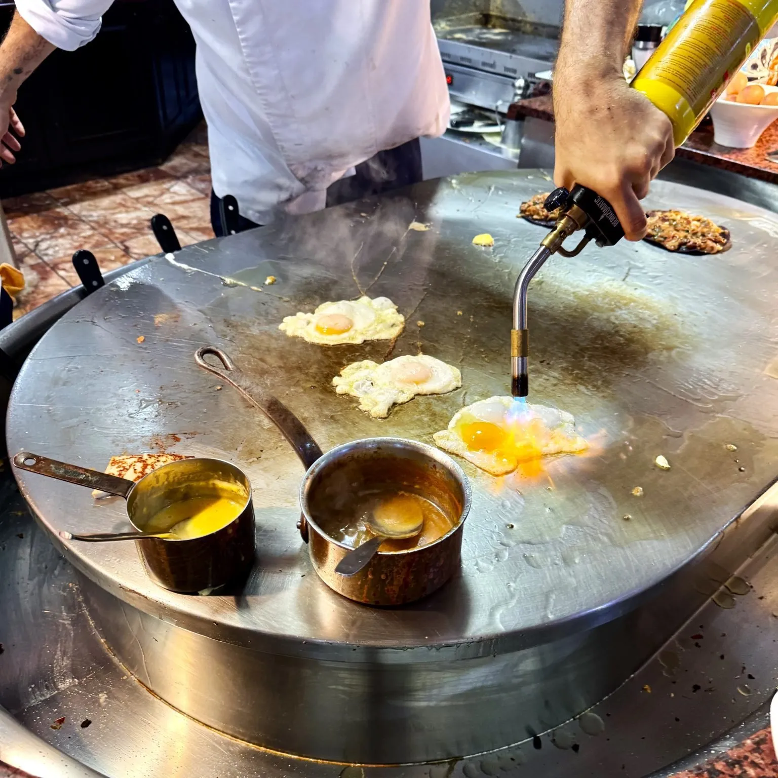 Cook using a blowtorch on fried eggs on the large circular chrome griddle