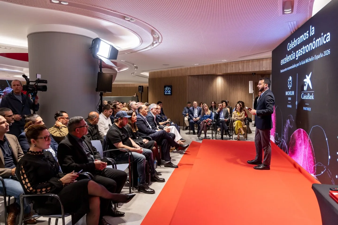 Vista general de la sala del acto: el presentador, de pie sobre la moqueta roja, se dirige al público sentado, con la pantalla que anuncia «Celebramos la excelencia gastronómica» al fondo