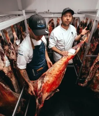 Two Taste 1973 cooks hold a goat carcass in the ageing chamber