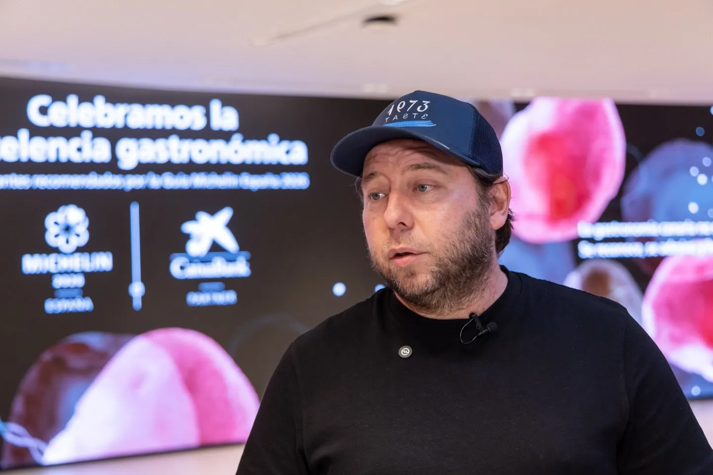 Retrato de Diego Schattenhofer con la gorra de Taste 1973, hablando frente a la pantalla del acto tras su intervención