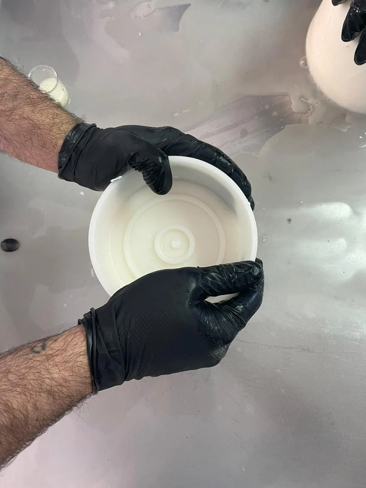 A gloved hand holds the lid of a cheese mould over the steel table