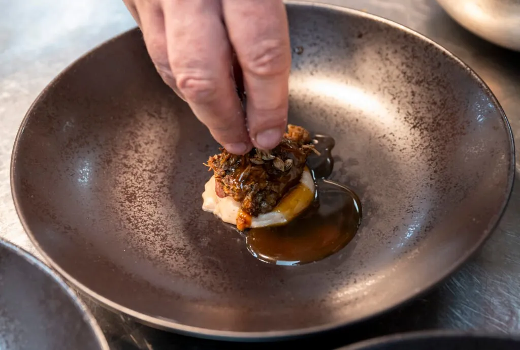 A chef finishes a dish by hand, served in a dark ceramic bowl