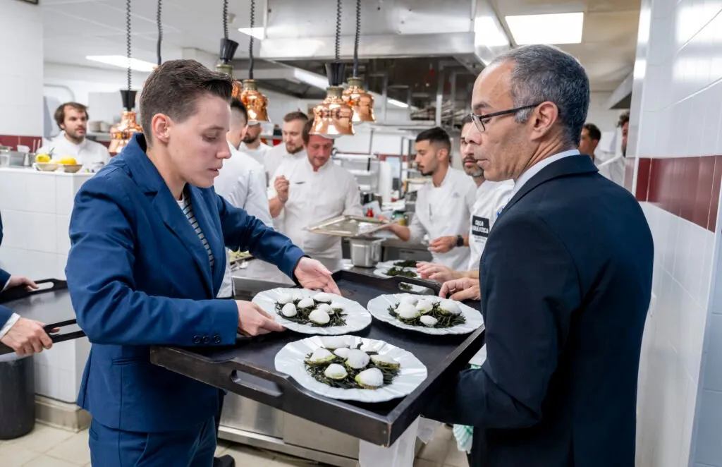 Two hosts bring trays of plated dishes out of the kitchen