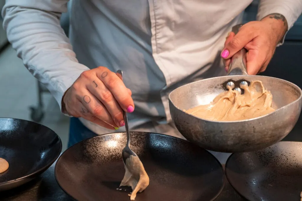 A chef’s hands plating a cream with a spoon onto deep plates