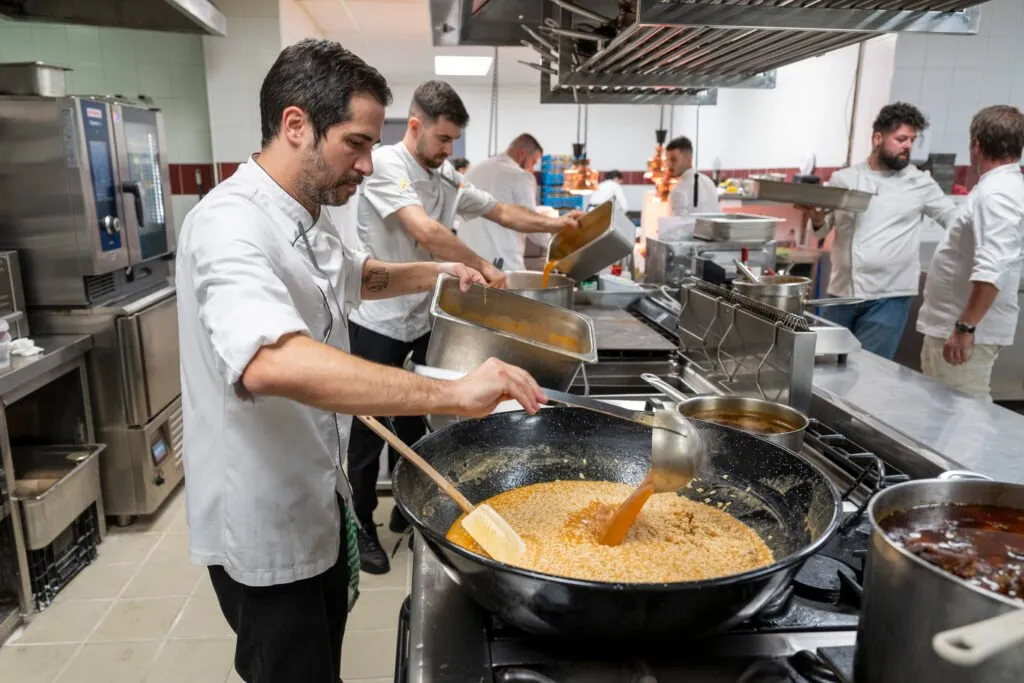 A chef pours stock with a ladle over a pan in the kitchen