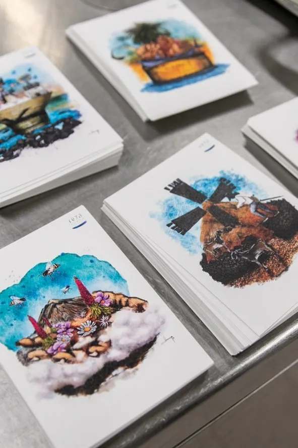 Watercolour cards showing dishes and island landscapes, stacked on the steel worktop of the kitchen
